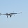 A fixed-wing unmanned aerial vehicle (UAV) is captured in flight against a clear blue sky. The aircraft features a long, slender fuselage with extended wings, retractable landing gear that appears partially deployed, and what appears to be sensor or payload equipment mounted underneath. The image is taken from a ground-level perspective looking up at the drone.