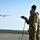 Two military personnel in camouflage uniforms are operating what appears to be a fixed-wing unmanned aerial vehicle (UAV/drone) from a runway or airstrip. One operator is wearing headset communications equipment and holding a control tablet mounted on a stand, while the UAV is visible in flight in the background. The setting appears to be a military airfield surrounded by forested terrain.