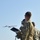Three military personnel in camouflage uniforms are observing a fixed-wing unmanned aerial vehicle (UAV) in flight. One soldier is wearing communications headset and holding what appears to be a control tablet or clipboard on a stand. The scene takes place on or near a tarmac/airstrip with a tree line visible in the background.