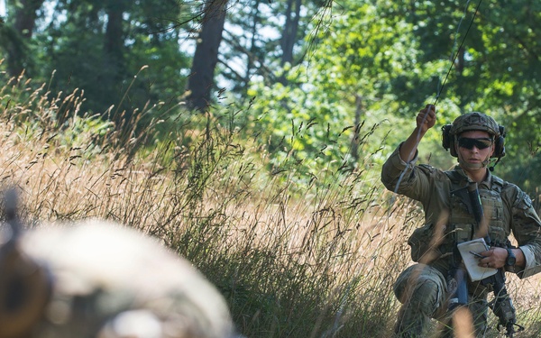 A soldier in camouflage uniform and helmet is kneeling in a grassy field near a tree line, holding a notepad and raising one hand, possibly giving a hand signal. In the foreground, another soldier's helmet and weapon are visible but out of focus. The setting appears to be a military field training exercise in a wooded, open terrain environment.