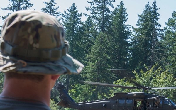 A military cadre member wearing a camouflage boonie hat and gray 'CADRE' t-shirt observes a UH-60 Black Hawk helicopter hovering low over an open grassy field. The helicopter appears to be conducting a training exercise, likely an air assault or personnel insertion/extraction operation. The setting is a rural clearing surrounded by dense coniferous forest, consistent with a Pacific Northwest training environment.