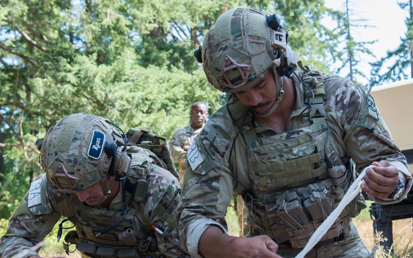 Two soldiers in U.S. Army MultiCam (OCP) uniforms are performing tactical combat casualty care (TCCC) on a training mannequin in an outdoor wooded environment. They appear to be applying bandages or tourniquets to the lower limbs of the simulated casualty. A third soldier observes in the background near a tan military vehicle, consistent with a training exercise.