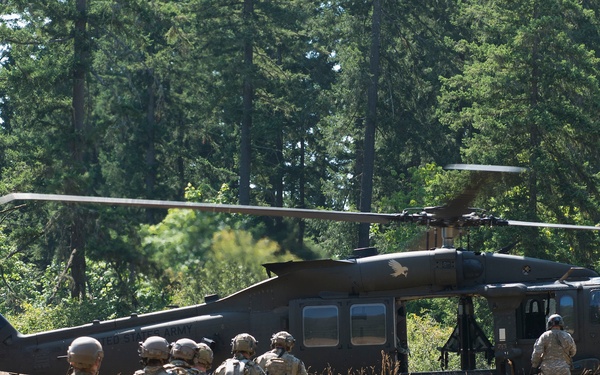 A group of military personnel in camouflage uniforms and helmets are walking across a dry grass field toward a dark olive-green military helicopter, consistent with a UH-60 Black Hawk. The scene appears to depict a troop insertion or extraction exercise in a wooded, open field environment. The helicopter is grounded with rotor blades visible and at least one crew member standing near the aircraft.