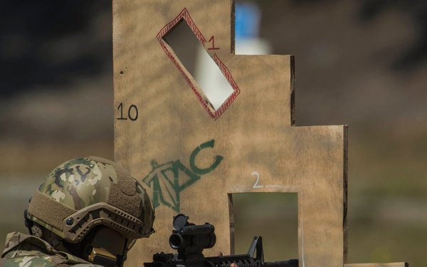 A soldier in full multicam camouflage gear and tactical helmet is firing an M4-style carbine rifle at a cardboard shooting range target. The target structure is a stepped cardboard silhouette with numbered zones (1, 2, 3) marked in different colors and labeled '5 ASOS' and '10'. A spent brass casing is visibly ejecting from the weapon during the firing sequence.