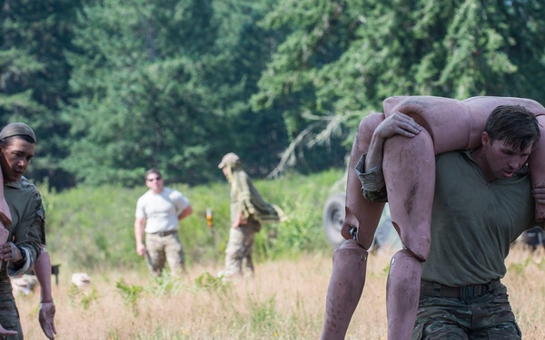 Military personnel in camouflage uniforms are conducting a casualty evacuation training exercise in an open field surrounded by trees. Two soldiers in the foreground are carrying training mannequins using a fireman's carry technique. Multiple other personnel and what appears to be an instructor or observer in a white shirt are visible in the background.