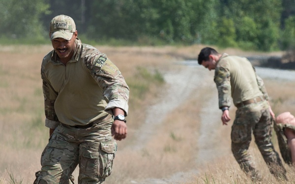 The image shows military personnel in camouflage uniforms conducting what appears to be a physical training or obstacle course exercise in an outdoor field setting. One uniformed individual is assisting or supervising a person in civilian clothing who is low to the ground, while a second uniformed individual performs a similar action in the background. The setting includes dry grassland, a dirt path, orange traffic cones, and tree line in the distance.