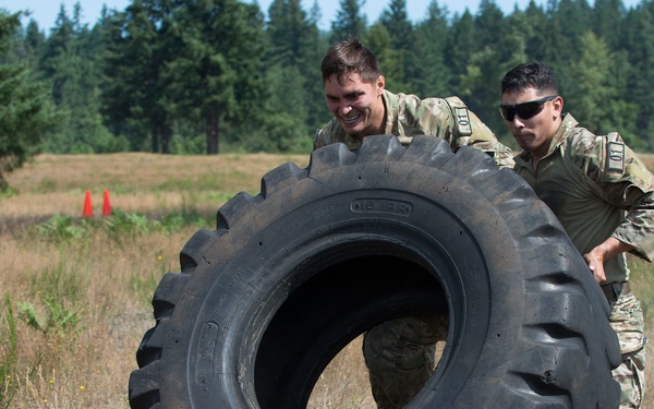 Two individuals in military camouflage uniforms are pushing a large heavy-duty tire across an open field. This appears to be a military physical training or obstacle course exercise conducted outdoors. Orange traffic cones are visible in the background, suggesting a marked course or training area.