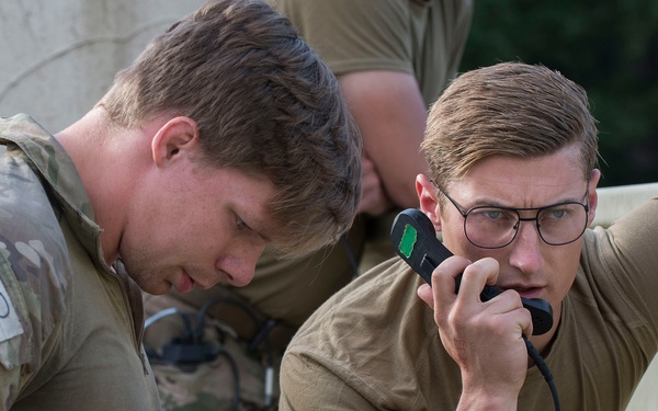 Two military personnel are engaged in what appears to be a communications exercise or operation. One soldier in full camouflage uniform is writing notes with a blue pen, while a second soldier in a olive/tan t-shirt speaks into a corded radio handset. A third individual is partially visible in the background.