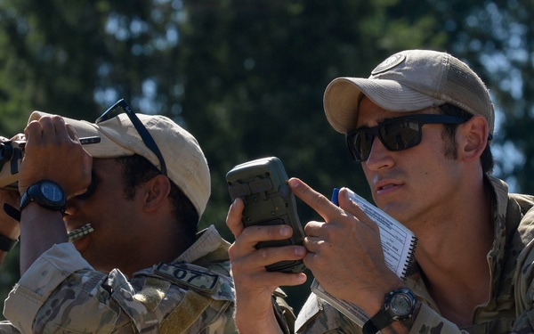 Two military personnel in multicam/OCP camouflage uniforms are conducting an observation or reconnaissance activity from an elevated position. One individual is using binoculars while the other holds a military-style device (likely a rangefinder or GPS unit) and a notepad with a pen. Both wear tan baseball caps, sunglasses, tactical vests, and wristwatches.