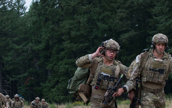 A group of military personnel in full combat gear are conducting a field exercise or patrol through an open grassy area with a dense conifer forest in the background. Two soldiers are prominently featured in the foreground, one adjusting communications equipment on their helmet while carrying a rifle, and another walking alongside with a weapon slung. Several additional soldiers are visible in the background following in a loose formation.