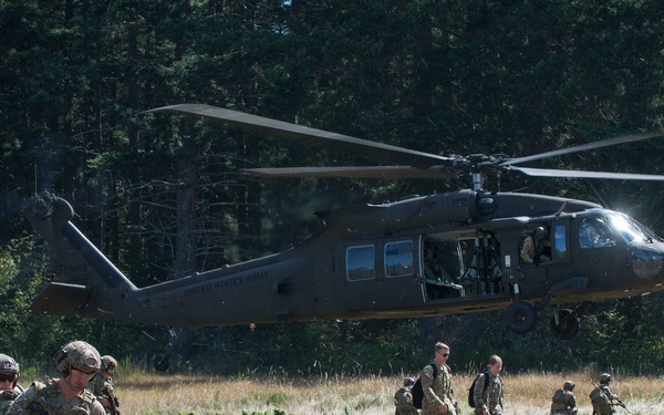 A military helicopter, identifiable as a UH-60 Black Hawk, hovers low over an open grassy field while multiple soldiers in full combat gear disembark or move away from the aircraft. The soldiers are wearing MultiCam/OCP camouflage uniforms, helmets, body armor, and carrying rifles and rucksacks. The scene appears to be a training exercise or tactical insertion operation conducted in a forested area.