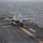 Marine F-35B Lightning II fighter aircraft complete GAU-22 cannon, ordnance hot reload exercise in Indo-Pacific Region An F-35B Lightning II stealth fighter jet is positioned on the flight deck of an amphibious assault ship, appearing to be preparing for or completing a short takeoff or vertical landing. The aircraft is centered on a yellow taxiway line with heat haze visible behind it, suggesting the engine is running at high power. The overcast sky and choppy ocean are visible in the background.