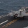 Marine F-35B Lightning II fighter aircraft complete GAU-22 cannon, ordnance hot reload exercise in Indo-Pacific Region An F-35B Lightning II stealth fighter jet is positioned on the flight deck of an amphibious assault ship at sea. The aircraft's lift fan door is open and the rear exhaust nozzle is visible in a downward-thrust configuration, consistent with Short Take-Off and Vertical Landing (STOVL) operations. The aircraft bears the number '01' and is photographed from an elevated angle with open ocean visible alongside the ship.
