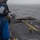 Marine F-35B Lightning II fighter aircraft complete GAU-22 cannon, ordnance hot reload exercise in Indo-Pacific Region A military jet aircraft, identifiable as an F-35B Lightning II, is positioned on the flight deck of a naval amphibious assault ship at sea. Deck crew members in color-coded safety gear are visible, with one signalman in yellow directing the aircraft. The overcast weather and open ocean backdrop suggest active at-sea operations.