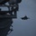 Marine F-35B Lightning II fighter aircraft complete GAU-22 cannon, ordnance hot reload exercise in Indo-Pacific Region A military jet aircraft is captured in flight against an overcast gray sky, with the superstructure of what appears to be a naval vessel visible in the blurred foreground. The aircraft has its landing gear deployed, suggesting it is either approaching for landing or has recently taken off. The low-light, overcast conditions and naval context suggest carrier-based flight operations.