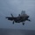 Marine F-35B Lightning II fighter aircraft complete GAU-22 cannon, ordnance hot reload exercise in Indo-Pacific Region An F-35B Lightning II stealth fighter jet is captured in a short takeoff or vertical landing (STOVL) configuration over open ocean, with landing gear deployed and lift fan door open. The aircraft appears to be operating from an amphibious assault ship or aircraft carrier, visible at the left edge of the frame. The image was taken under overcast, low-light conditions over a gray sea.