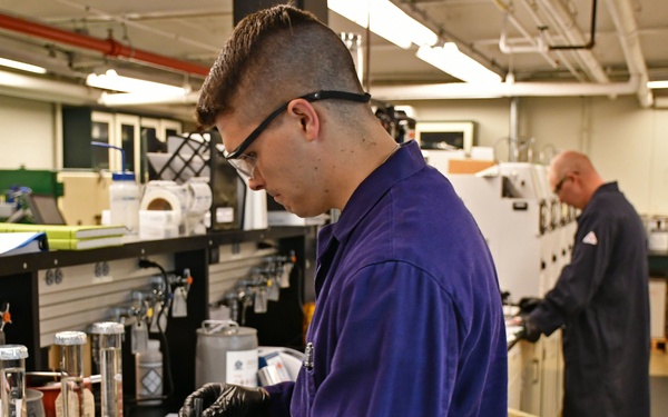 Two individuals are working in a professional laboratory setting. The foreground technician, wearing a purple lab coat, safety glasses, and black nitrile gloves, is handling small instruments or samples at a workbench. A second person in the background, wearing military camouflage pants and a dark jacket, is working at a separate station with analytical equipment.