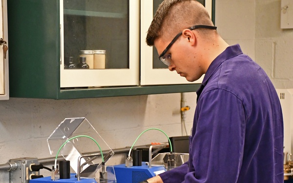 A young male laboratory technician wearing a purple lab coat, safety glasses, and black nitrile gloves is operating a blue PAC PMD 110 analytical instrument in a laboratory setting. The lab is well-equipped with various instruments, storage cabinets, and chemical containers. The setting appears to be an industrial or materials testing laboratory.