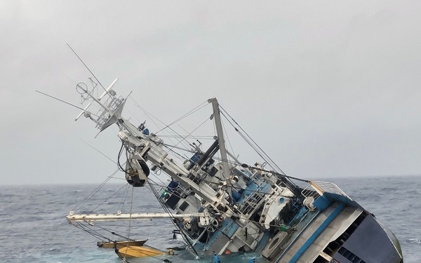 A large fishing vessel is capsizing and sinking at sea, listing heavily to starboard at an extreme angle. Fishing gear including extensive yellow float lines and netting has spilled into the surrounding water. An orange inflatable life raft and a blue rigid rescue craft are visible in the water near the stricken vessel, indicating an emergency evacuation situation.