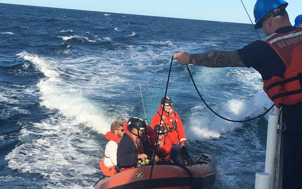 A U.S. Coast Guard operation is underway at sea, showing personnel in an orange inflatable rescue boat (RIB) being tethered or guided alongside a larger Coast Guard vessel. Several individuals wearing orange life vests, helmets, and safety gear are visible both on the small boat and on the deck of the larger ship. The sea conditions appear moderately rough with visible wave action and white water around the vessels.
