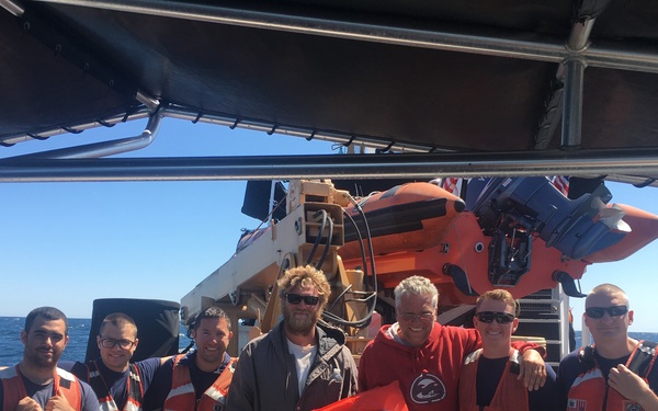 A group of seven people pose together on the deck of what appears to be a Coast Guard vessel at sea. Five individuals are wearing orange Coast Guard life vests over dark uniforms, while two individuals in the center appear to be civilians, possibly rescued persons. The photo is taken on a clear, sunny day with calm blue ocean visible in the background.