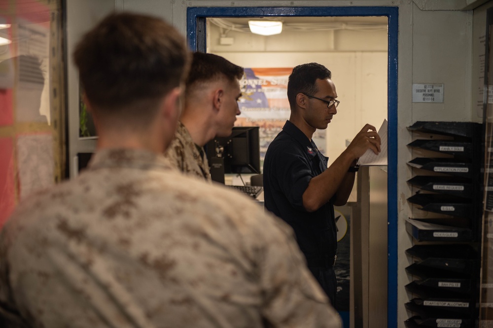 The image shows military personnel in what appears to be an administrative office aboard a naval vessel or military installation. A Navy sailor in a dark uniform is reviewing a document at a filing station, while two Marines in camouflage uniforms wait in the foreground. The setting appears to be a personnel services or administrative office based on visible signage and document organizers.