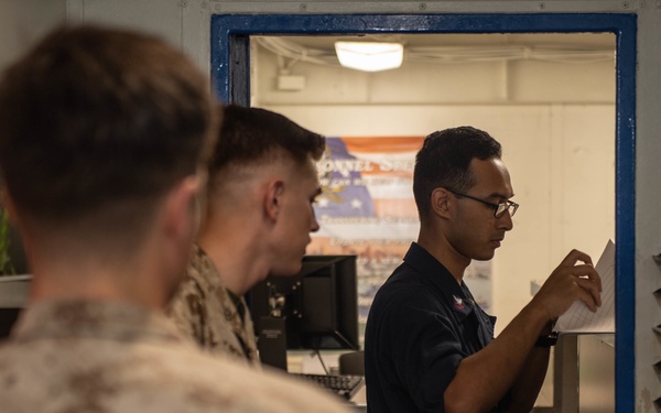 The image shows military personnel in what appears to be an administrative office aboard a naval vessel or military installation. A Navy sailor in a dark uniform is reviewing a document at a filing station, while two Marines in camouflage uniforms wait in the foreground. The setting appears to be a personnel services or administrative office based on visible signage and document organizers.