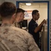 The image shows military personnel in what appears to be an administrative office aboard a naval vessel or military installation. A Navy sailor in a dark uniform is reviewing a document at a filing station, while two Marines in camouflage uniforms wait in the foreground. The setting appears to be a personnel services or administrative office based on visible signage and document organizers.