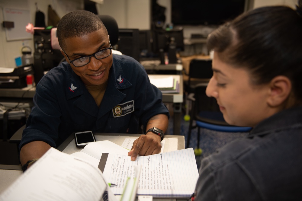Two individuals in a military or naval setting are engaged in a study or review session, with one person in a Navy uniform pointing at handwritten notes in an open notebook. The uniformed individual appears to be explaining or instructing the other person, who is listening attentively. The setting appears to be an office or administrative workspace aboard a naval vessel or installation.