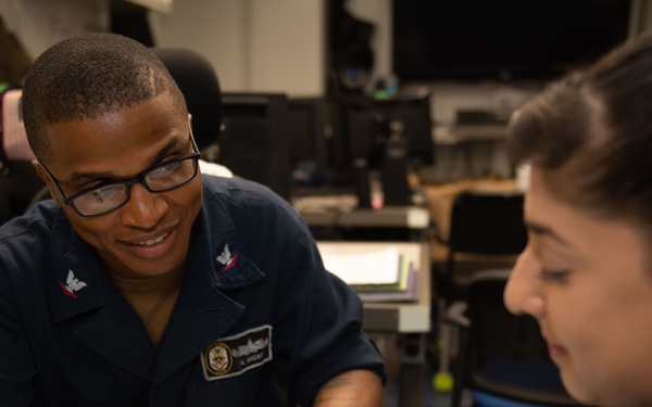 Two individuals in a military or naval setting are engaged in a study or review session, with one person in a Navy uniform pointing at handwritten notes in an open notebook. The uniformed individual appears to be explaining or instructing the other person, who is listening attentively. The setting appears to be an office or administrative workspace aboard a naval vessel or installation.