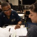 Two individuals in a military or naval setting are engaged in a study or review session, with one person in a Navy uniform pointing at handwritten notes in an open notebook. The uniformed individual appears to be explaining or instructing the other person, who is listening attentively. The setting appears to be an office or administrative workspace aboard a naval vessel or installation.