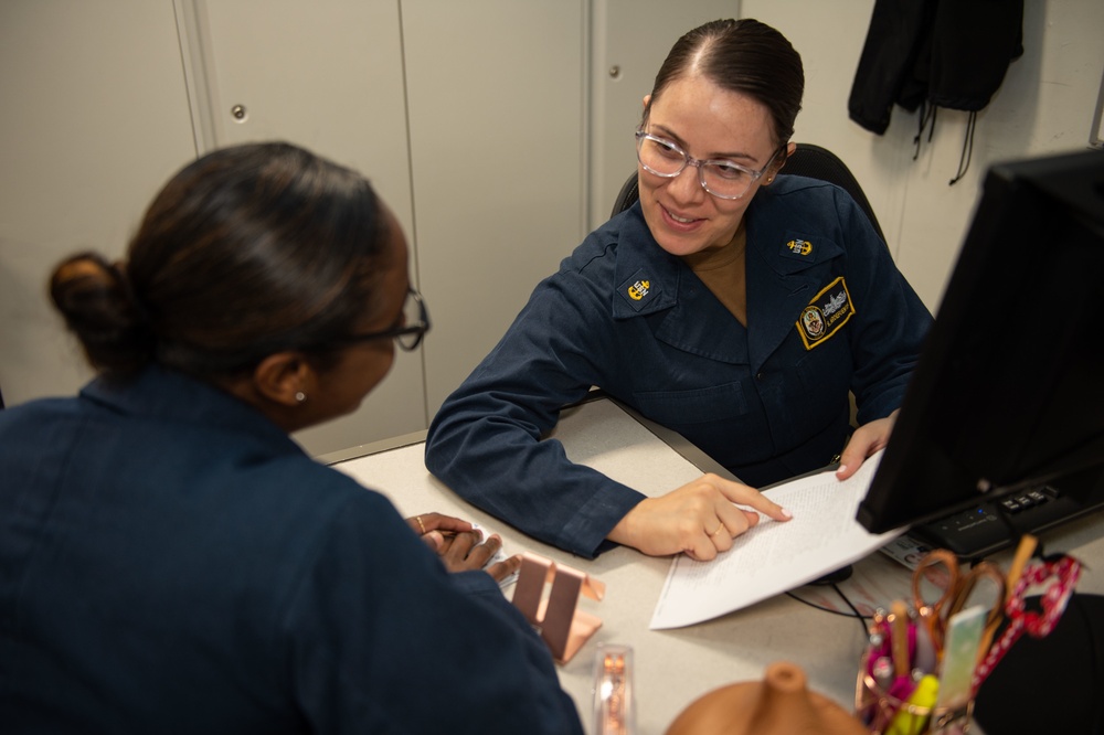 Two individuals wearing matching dark navy blue military uniforms are seated at a desk in what appears to be an office or administrative workspace. The person facing the camera is smiling and pointing to a document, appearing to explain or review its contents with the other individual. The setting suggests a formal military administrative or counseling interaction.