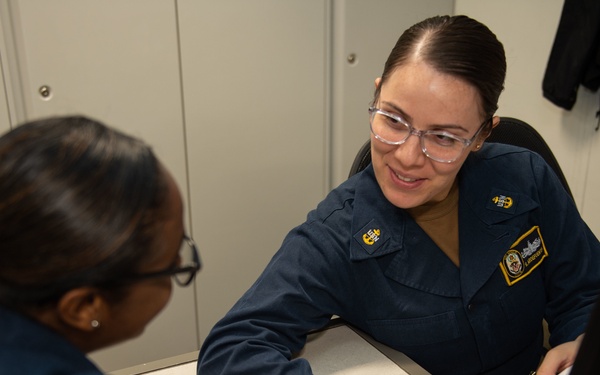 Two individuals wearing matching dark navy blue military uniforms are seated at a desk in what appears to be an office or administrative workspace. The person facing the camera is smiling and pointing to a document, appearing to explain or review its contents with the other individual. The setting suggests a formal military administrative or counseling interaction.