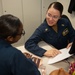 Two individuals wearing matching dark navy blue military uniforms are seated at a desk in what appears to be an office or administrative workspace. The person facing the camera is smiling and pointing to a document, appearing to explain or review its contents with the other individual. The setting suggests a formal military administrative or counseling interaction.