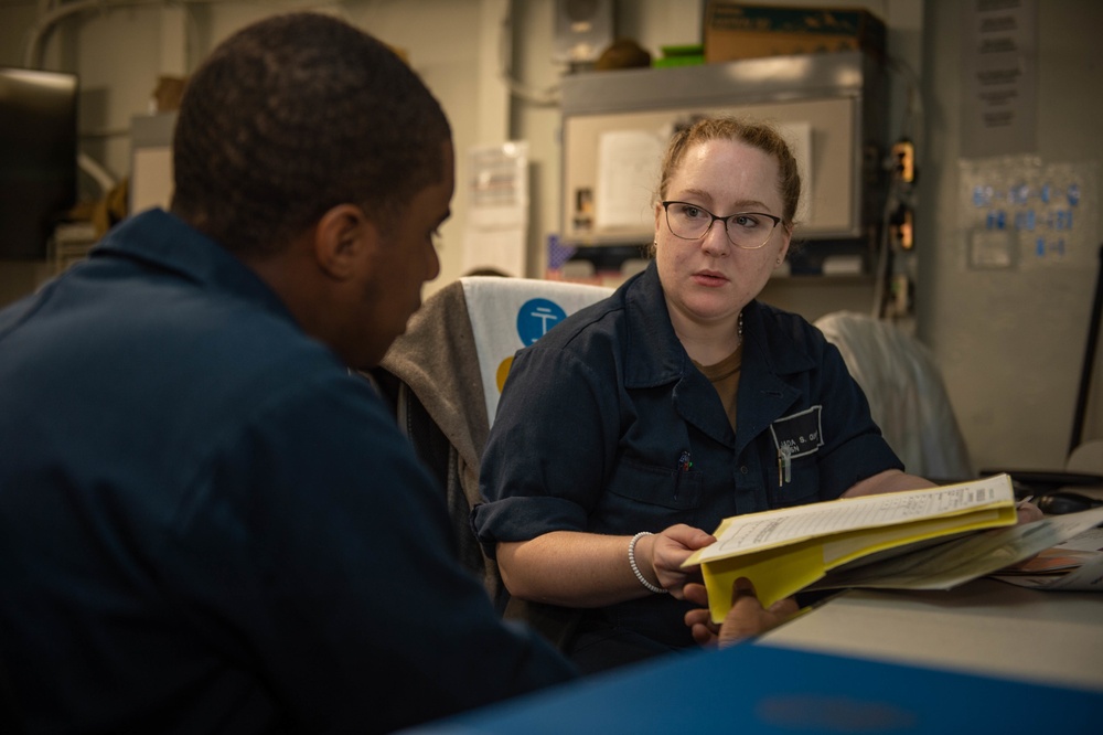 Two individuals wearing matching navy blue uniforms are engaged in a work-related discussion in what appears to be a military or naval workspace. The person facing the camera, a woman wearing glasses, is holding an open yellow binder and appears to be reviewing or explaining its contents. The setting features institutional gray walls with mounted equipment and posted documents.