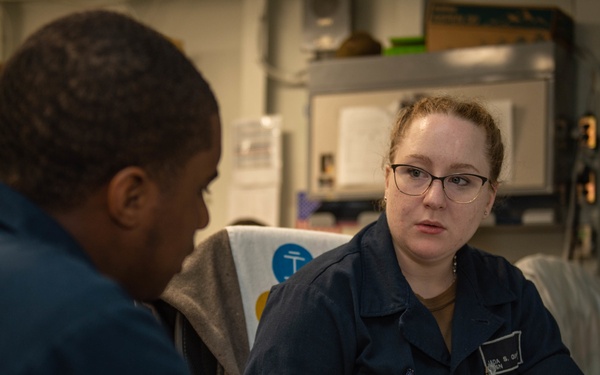 Two individuals wearing matching navy blue uniforms are engaged in a work-related discussion in what appears to be a military or naval workspace. The person facing the camera, a woman wearing glasses, is holding an open yellow binder and appears to be reviewing or explaining its contents. The setting features institutional gray walls with mounted equipment and posted documents.