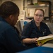 Two individuals wearing matching navy blue uniforms are engaged in a work-related discussion in what appears to be a military or naval workspace. The person facing the camera, a woman wearing glasses, is holding an open yellow binder and appears to be reviewing or explaining its contents. The setting features institutional gray walls with mounted equipment and posted documents.