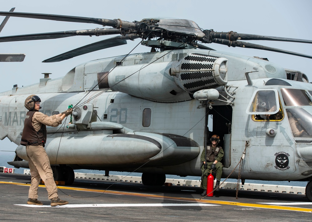 A large military helicopter, identified as a CH-53E Super Stallion bearing 'MAR' and number '20' markings, is positioned on what appears to be a naval vessel flight deck. Two personnel in flight gear are visible — one standing and handling rotor tie-down lines, and another seated in the open side door. A red fire extinguisher is positioned near the aircraft as a safety precaution.
