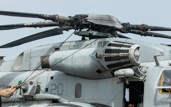 A large military helicopter, identified as a CH-53E Super Stallion bearing 'MAR' and number '20' markings, is positioned on what appears to be a naval vessel flight deck. Two personnel in flight gear are visible — one standing and handling rotor tie-down lines, and another seated in the open side door. A red fire extinguisher is positioned near the aircraft as a safety precaution.