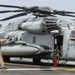 A large military helicopter, identified as a CH-53E Super Stallion bearing 'MAR' and number '20' markings, is positioned on what appears to be a naval vessel flight deck. Two personnel in flight gear are visible — one standing and handling rotor tie-down lines, and another seated in the open side door. A red fire extinguisher is positioned near the aircraft as a safety precaution.