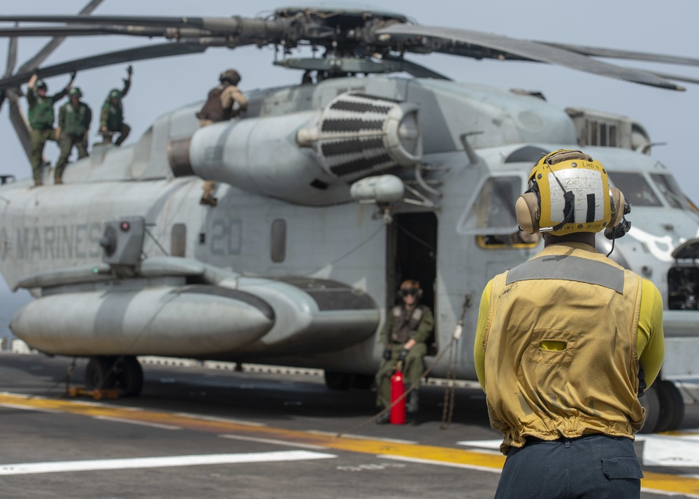 A U.S. Marines helicopter, identified as a CH-53E Super Stallion (number 20), is positioned on what appears to be a naval vessel's flight deck. A deck crew member in a yellow jersey and cranial helmet stands in the foreground observing the aircraft, while maintenance or flight crew personnel are visible on top of and inside the helicopter. The scene depicts pre-flight or maintenance operations on a ship's flight deck.