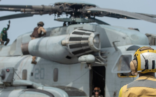 A U.S. Marines helicopter, identified as a CH-53E Super Stallion (number 20), is positioned on what appears to be a naval vessel's flight deck. A deck crew member in a yellow jersey and cranial helmet stands in the foreground observing the aircraft, while maintenance or flight crew personnel are visible on top of and inside the helicopter. The scene depicts pre-flight or maintenance operations on a ship's flight deck.