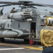 A U.S. Marines helicopter, identified as a CH-53E Super Stallion (number 20), is positioned on what appears to be a naval vessel's flight deck. A deck crew member in a yellow jersey and cranial helmet stands in the foreground observing the aircraft, while maintenance or flight crew personnel are visible on top of and inside the helicopter. The scene depicts pre-flight or maintenance operations on a ship's flight deck.