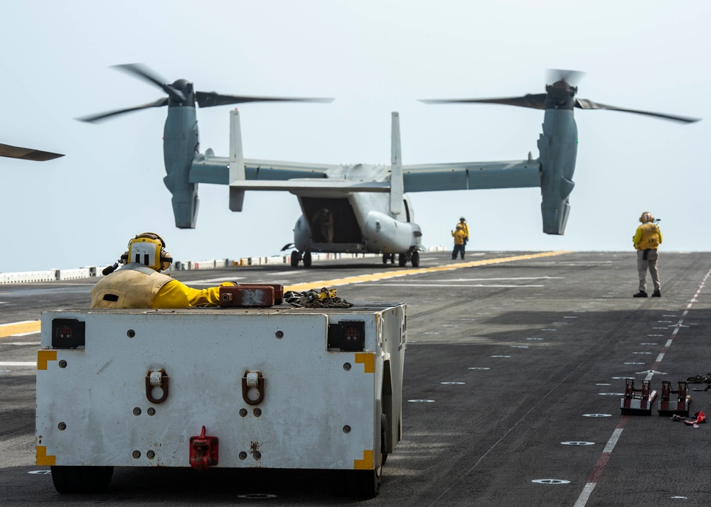 A V-22 Osprey tiltrotor aircraft is on the flight deck of a naval vessel, with its rotors spinning in helicopter mode. Several deck crew members in yellow jerseys are visible performing flight deck operations. Ground support equipment, including a deck tractor/mule, is positioned in the foreground.
