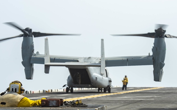 A V-22 Osprey tiltrotor aircraft is on the flight deck of a naval vessel, with its rotors spinning in helicopter mode. Several deck crew members in yellow jerseys are visible performing flight deck operations. Ground support equipment, including a deck tractor/mule, is positioned in the foreground.