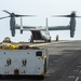 A V-22 Osprey tiltrotor aircraft is on the flight deck of a naval vessel, with its rotors spinning in helicopter mode. Several deck crew members in yellow jerseys are visible performing flight deck operations. Ground support equipment, including a deck tractor/mule, is positioned in the foreground.