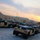 A large motor pool of desert-tan HMMWV (Humvee) military vehicles parked in organized rows on a hardstand surface at what appears to be a forward operating base or military installation. The vehicles appear to be open-top configurations with cargo/equipment covered by dark tarps or netting. The scene is photographed at sunset or sunrise, casting warm orange and yellow tones across the sky.