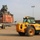 A military logistics yard showing heavy equipment operations with a Terex reach stacker lifting a red shipping container marked with US Army markings. A yellow JCB 535-140 telehandler is positioned in the foreground on a dusty, arid terrain. A worker in military-style clothing observes the container lift operation in the background.