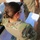 A U.S. Army soldier named NISBET is reviewing documents on a clipboard in what appears to be an outdoor logistics or supply operation. She is wearing OCP (Operational Camouflage Pattern) uniform with a subdued American flag patch on her right sleeve. In the background, there are supply boxes including one labeled with 'FRAGO 19-1221 BINOCULARS' with a quantity of 17.
