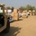 The image shows a military logistics or humanitarian aid distribution operation in an arid environment. Multiple pallets stacked with cardboard boxes and labeled supplies, including items marked 'SURE-Pak', are lined up on a dirt surface. Military personnel in desert camouflage uniforms and military vehicles, including a green/tan HMMWV (Humvee), are present at the site.